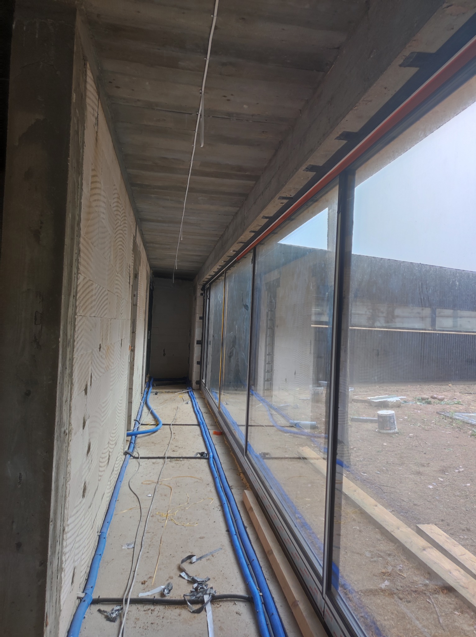 Wnętrze surowego budynku w trakcie prac wykończeniowych, widoczne ściany z bloczków, betonowy strop, okno panoramiczne z widokiem na zewnątrz, niebieskie rury instalacyjne prowadzone wzdłuż ściany.