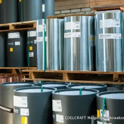 Magazyn z rolkami blachy w odcieniach szarości, srebrnej i miedzianej, ułożonymi na drewnianych paletach. Widoczne etykiety producenta.