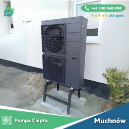 Energia Taniej Sp. z o.o. - Szara, dwuczęściowa jednostka zewnętrzna z wentylatorami na metalowym stelażu, ustawiona na betonowej podstawie przy białej ścianie budynku z widocznym oknem. W tle zielony krzew.