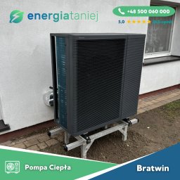 Energia Taniej Sp. z o.o. - Czarna pompa ciepła Panasonic Aquarea na srebrnej konstrukcji wsporczej, zamontowana przy białej ścianie budynku z widocznym oknem i fragmentem chodnika z kostki.