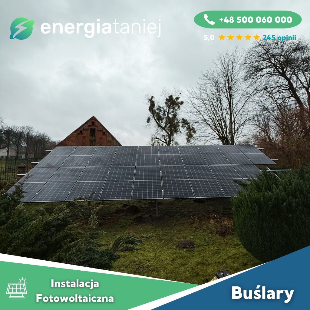 Instalacja fotowoltaiczna na gruncie przed ceglanym budynkiem gospodarczym w pochmurny dzień, widoczne krzewy i drzewa bez liści.