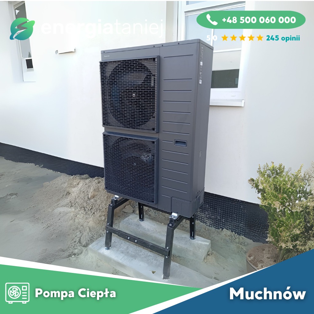 Szara, dwuczęściowa jednostka zewnętrzna z wentylatorami na metalowym stelażu, ustawiona na betonowej podstawie przy białej ścianie budynku z widocznym oknem. W tle zielony krzew.