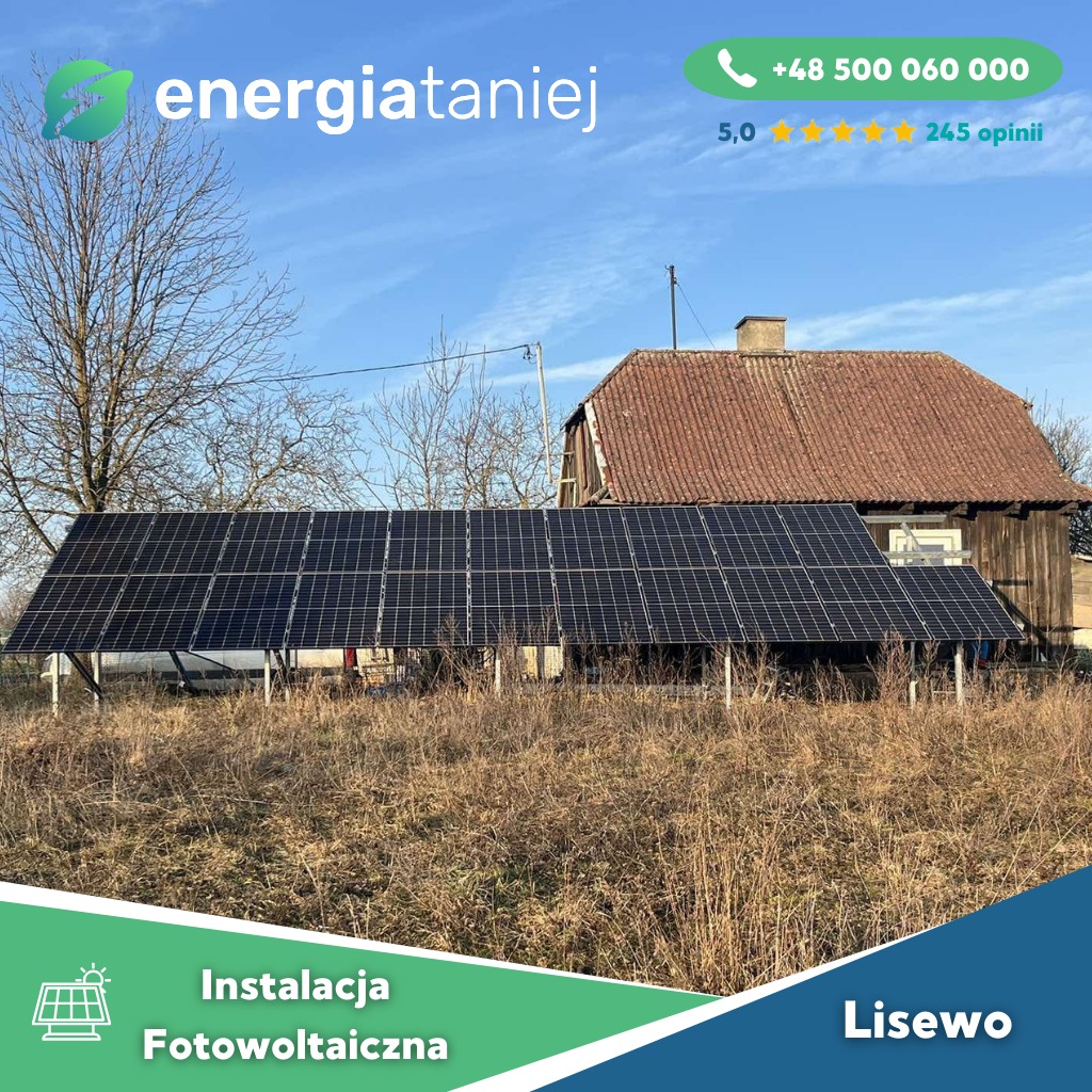 Panel słoneczny zamontowany na konstrukcji na tle starego, ceglanego budynku gospodarczego i bezlistnego drzewa pod błękitnym niebem.