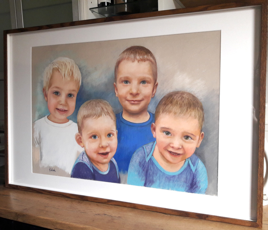 Portret pastelowy czterech uśmiechniętych chłopców w ramie z brązowego drewna, widoczny z boku, na drewnianym blacie.