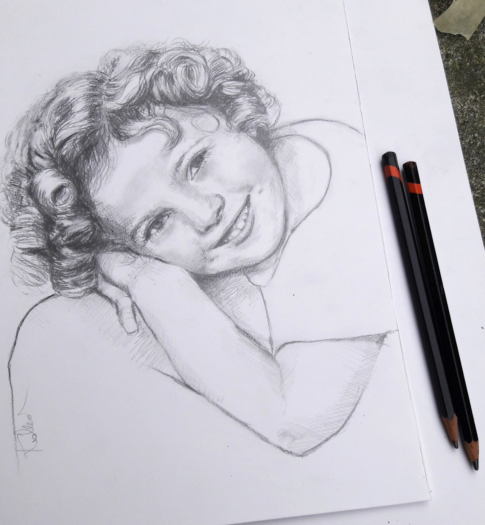 Szkic ołówkiem przedstawiający portret uśmiechniętej dziewczynki z kręconymi włosami, obok leżą dwa ołówki z czerwonymi paskami.