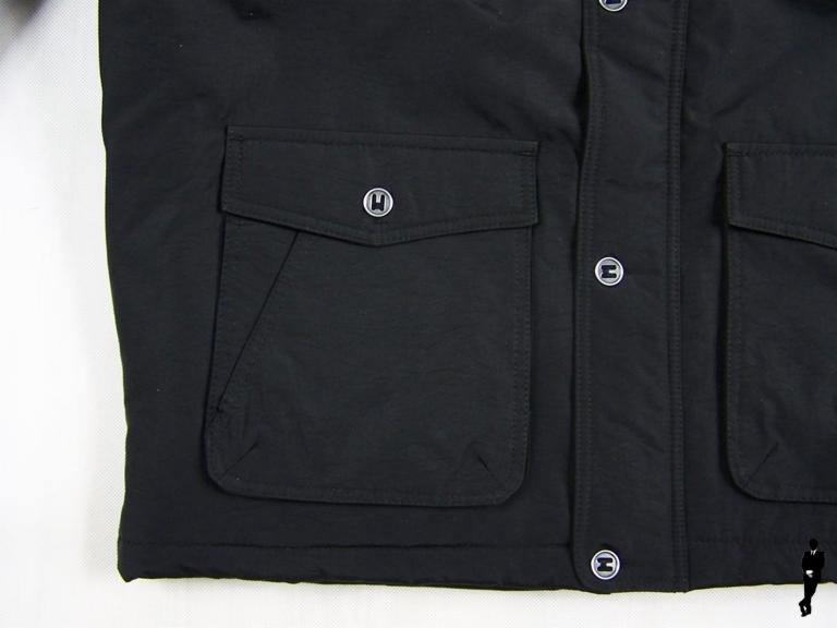 Czarna kurtka z kieszeniami na guziki, zbliżenie na detal wykończenia i fakturę materiału. Widoczny fragment wszywki i guziki z logo.