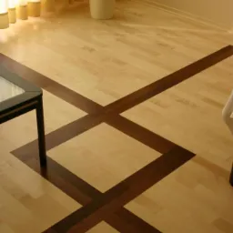 Jasna drewniana podłoga z intarsją z ciemniejszego drewna tworzącą geometryczny wzór krzyża i kwadratu, widoczny fragment stolika kawowego z czarnymi nogami, roślina doniczkowa w tle.