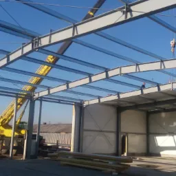 Wnętrze hali w trakcie budowy, widoczne stalowe elementy konstrukcyjne, żółty dźwig na zewnątrz i pracownik na dachu przy świetle dziennym.