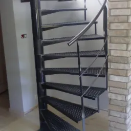 Czarna, metalowa, spiralna klatka schodowa z perforowanymi stopniami, umieszczona w jasnym wnętrzu obok ceglanej ściany. Widoczne spawane elementy konstrukcji.