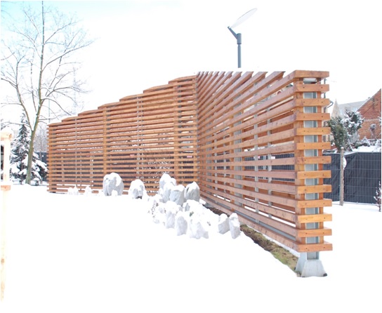 Drewniana, pozioma, ażurowa konstrukcja ogrodzenia z metalowymi słupkami na tle zimowego krajobrazu. Elementy konstrukcyjne w regularnych odstępach.