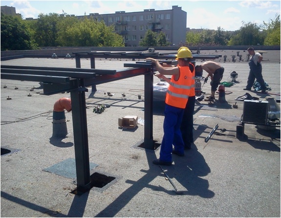 Montaż metalowej konstrukcji na dachu budynku przez robotników w kaskach i kamizelkach odblaskowych. Widoczne narzędzia i elementy konstrukcyjne.