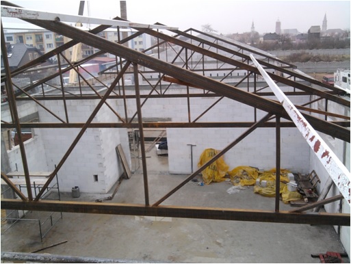 Konstrukcja stalowa dachu budynku w trakcie budowy, widok z góry. Surowe ściany z betonu komórkowego, w tle zabudowa miejska i kościół.