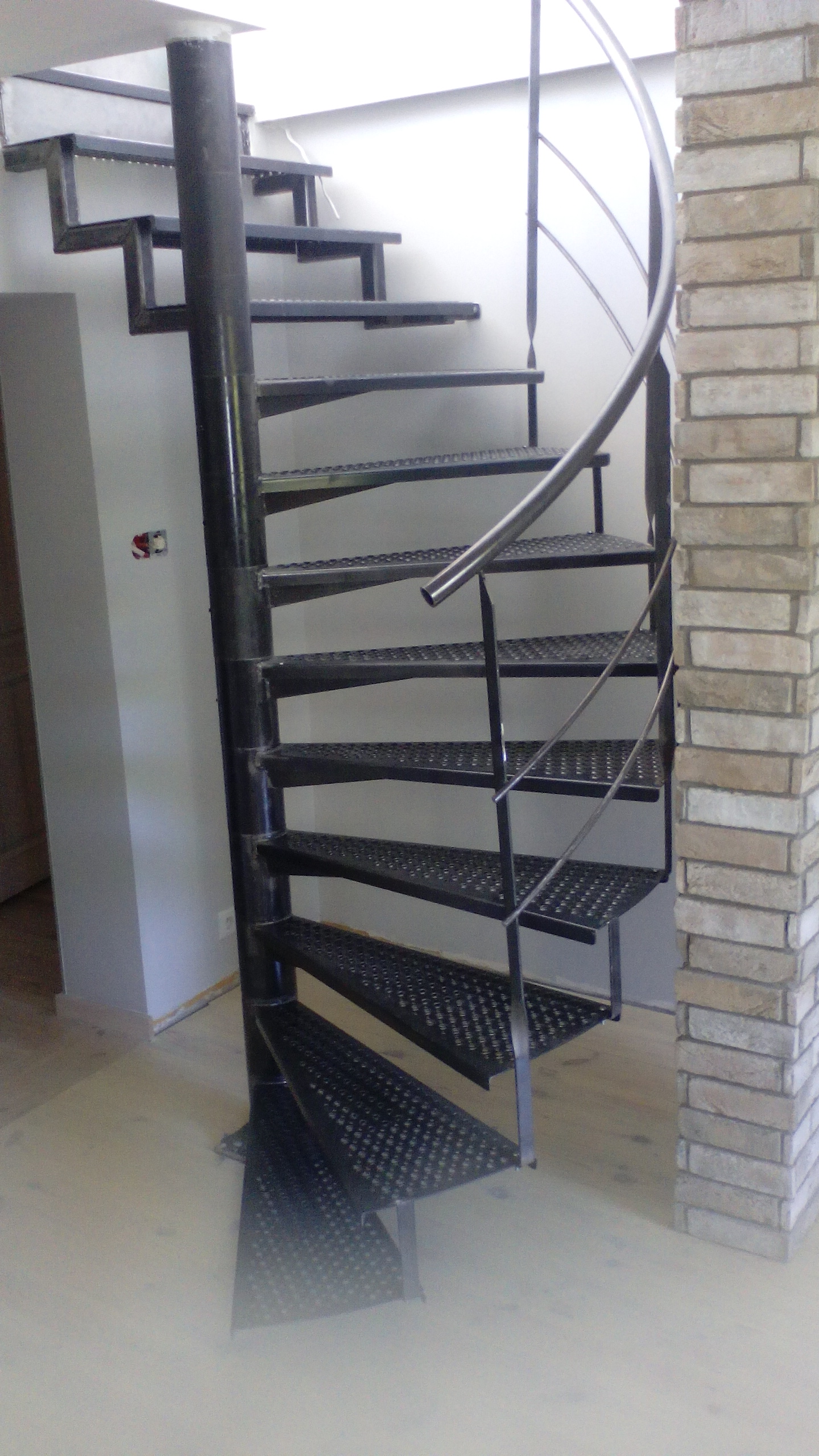 Czarna, metalowa, spiralna klatka schodowa z perforowanymi stopniami, umieszczona w jasnym wnętrzu obok ceglanej ściany. Widoczne spawane elementy konstrukcji.