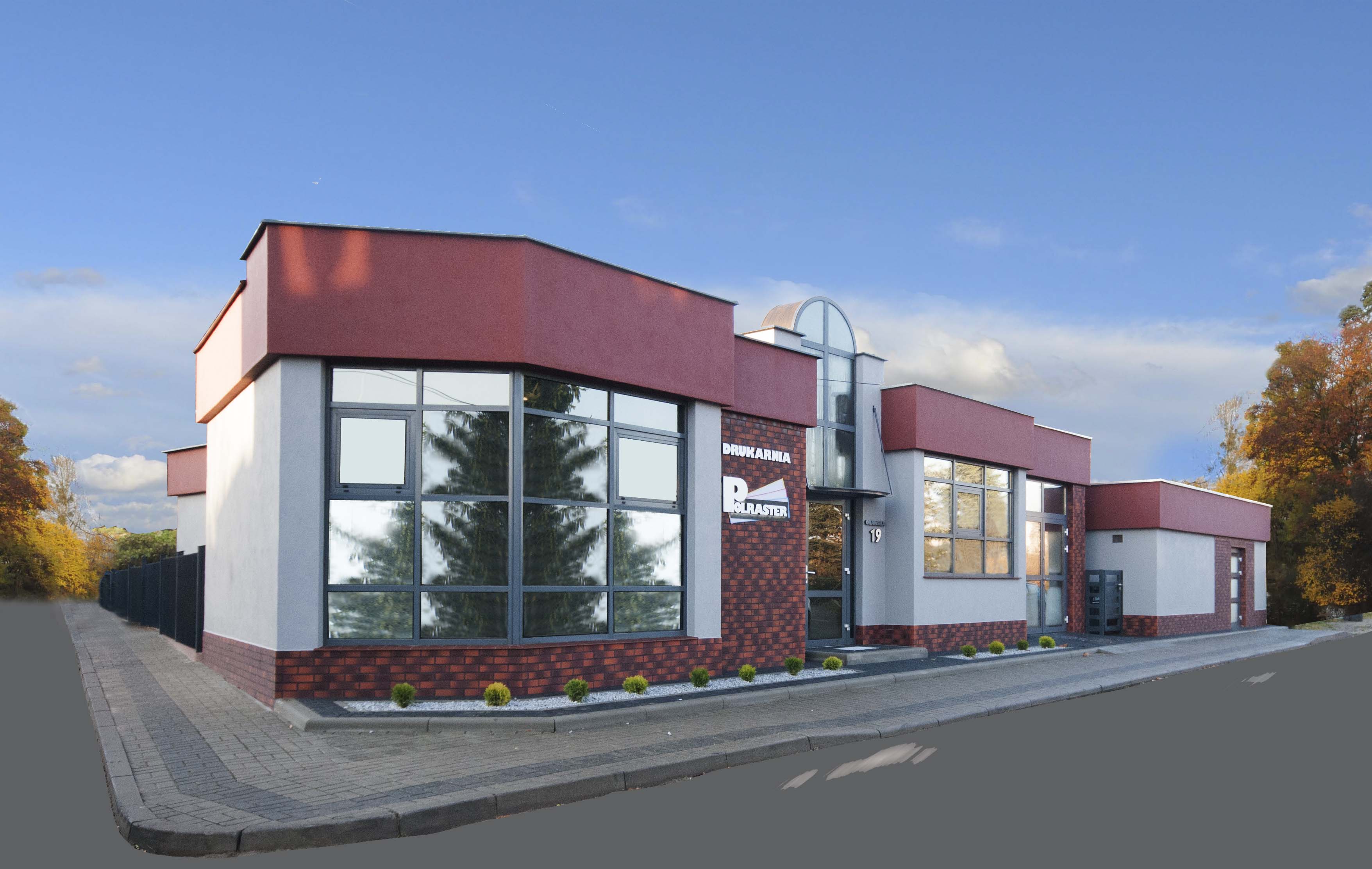 Budynek drukarni Polraster z dużymi oknami, ceglaną fasadą i logo firmy. Widok na ulicę i drzewa w tle, błękitne niebo.