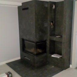 Tech-Kras P.H.U. - Nowoczesny kominek obłożony ciemnym kamieniem z półkami, widoczny w rogu pomieszczenia z jasnymi ścianami i podłogą. Minimalistyczny design.