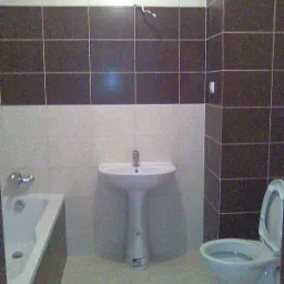 Wykończona łazienka z brązowo-białymi płytkami, białą umywalką na postumencie, toaletą i fragmentem wanny. Widoczny kran i odpływ w wannie.