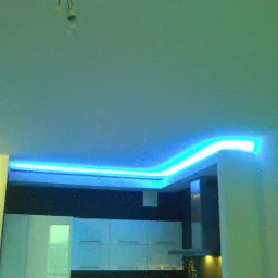 Niebieskie, neonowe oświetlenie sufitowe w nowoczesnym wnętrzu. Widoczna część kuchni z białymi szafkami i okapem. W tle ciemna ściana.