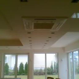 Podwieszany sufit z klimatyzacją i oświetleniem punktowym w jasnym pomieszczeniu z dużymi oknami i widokiem na zieleń. Nowoczesne wykończenie wnętrza.