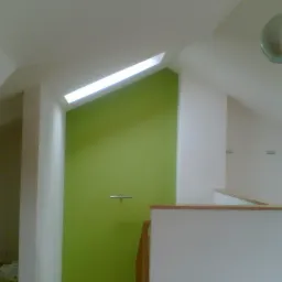 Ściana pomalowana na zielono z oknem dachowym i elementami wykończenia w jasnym wnętrzu poddasza, widok na fragment balustrady i lampę.