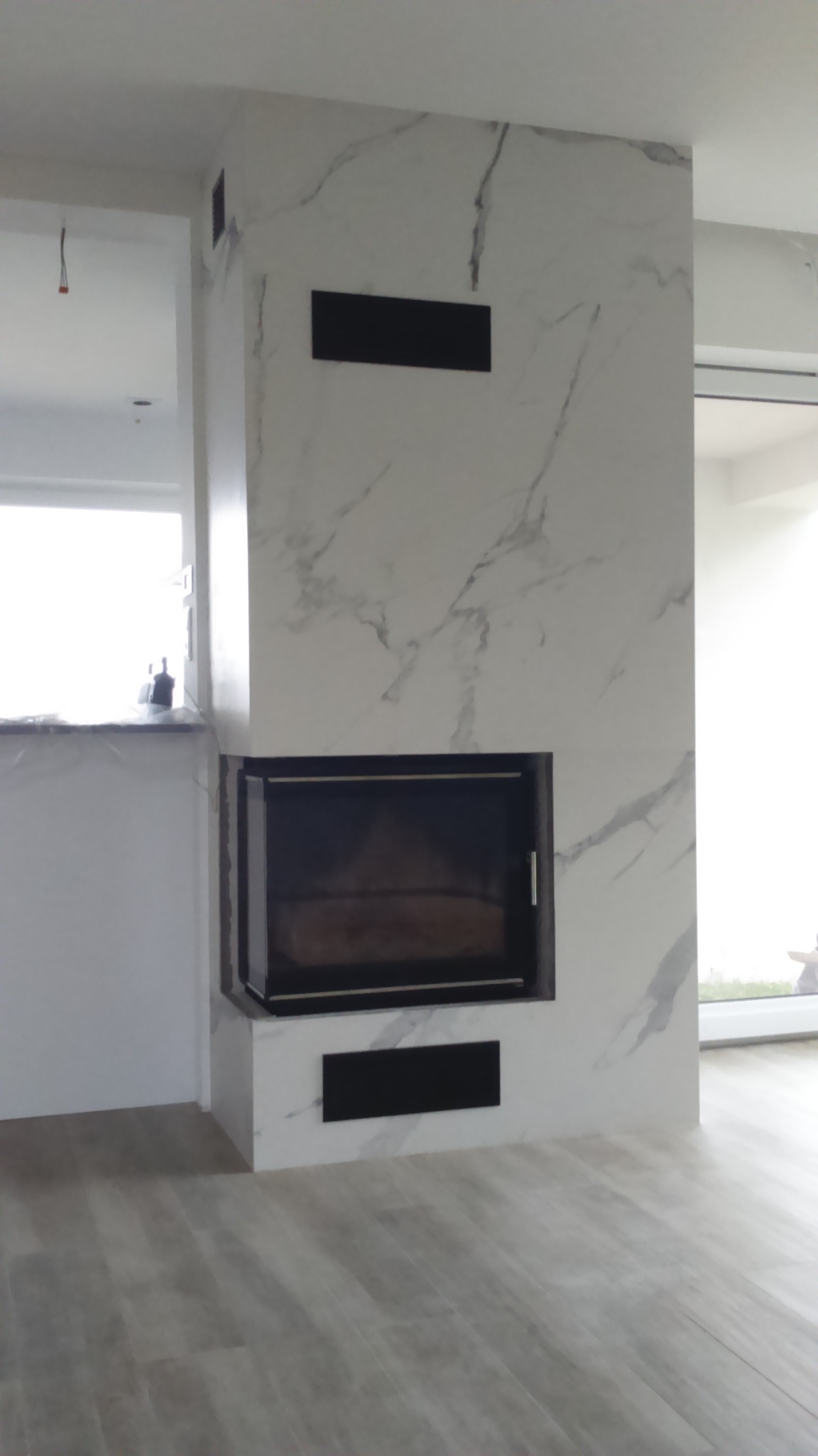 Obudowa kominka z płyt imitujących marmur, z czarnymi wstawkami wentylacyjnymi. Jasna podłoga z paneli. Nowoczesny design, wykończenie wnętrza.