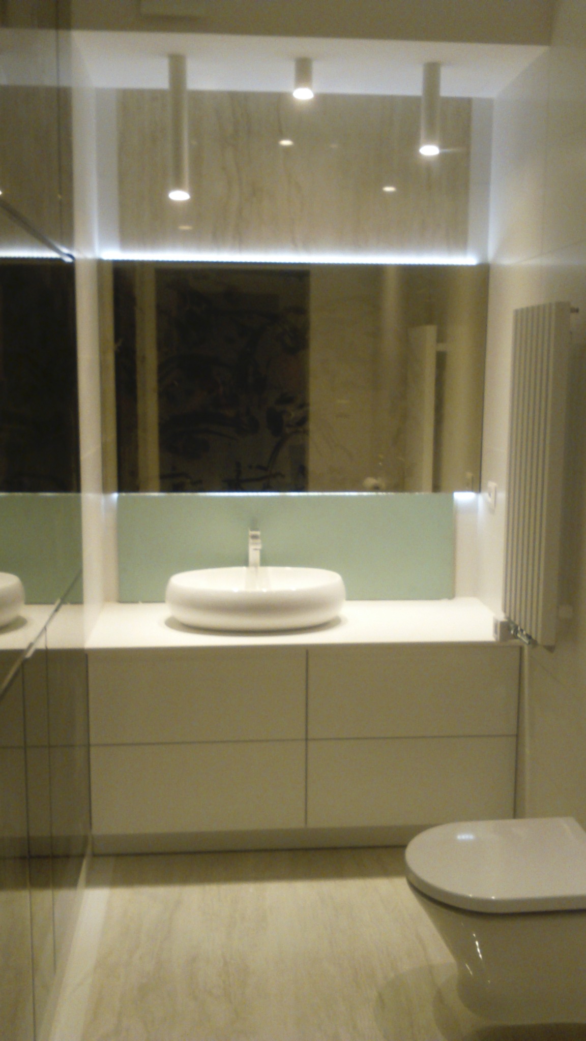 Nowoczesna łazienka z owalną umywalką na szafce, podświetlanym lustrem i grzejnikiem drabinkowym. Minimalistyczny design, jasne kolory, punktowe oświetlenie sufitowe.