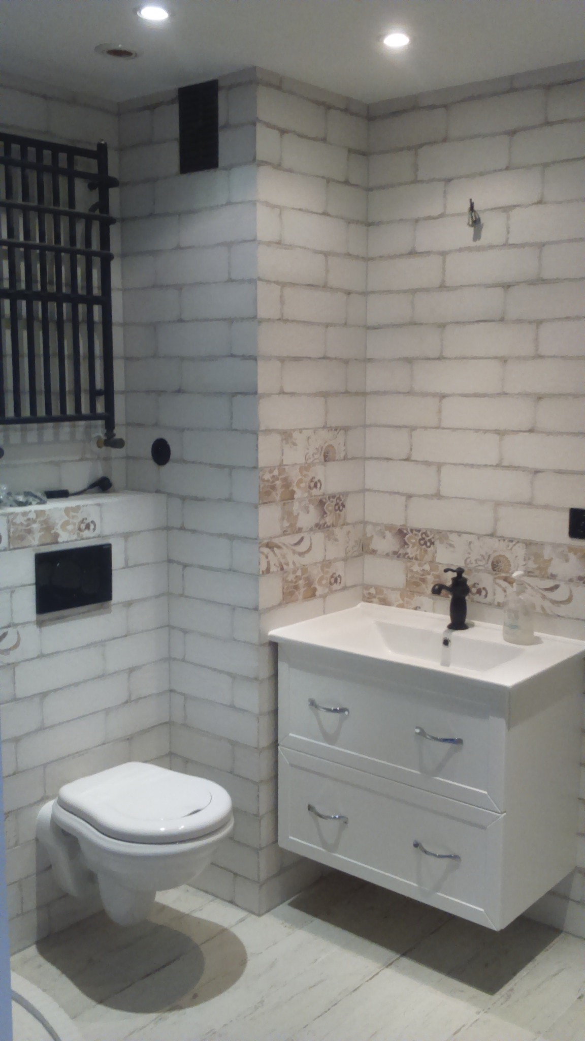 Wykończona łazienka z białymi płytkami imitującymi cegłę, wiszącą toaletą i szafką pod umywalką. Dekoracyjne płytki z motywem kwiatowym. Grzejnik drabinkowy.