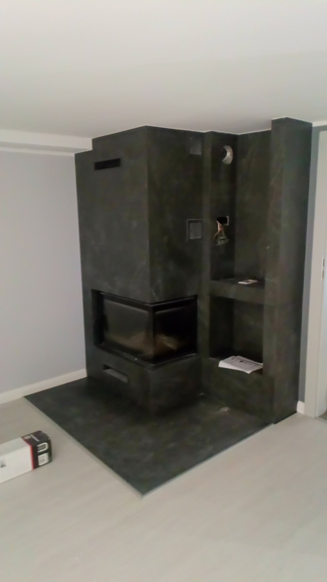 Nowoczesny kominek obłożony ciemnym kamieniem z półkami, widoczny w rogu pomieszczenia z jasnymi ścianami i podłogą. Minimalistyczny design.