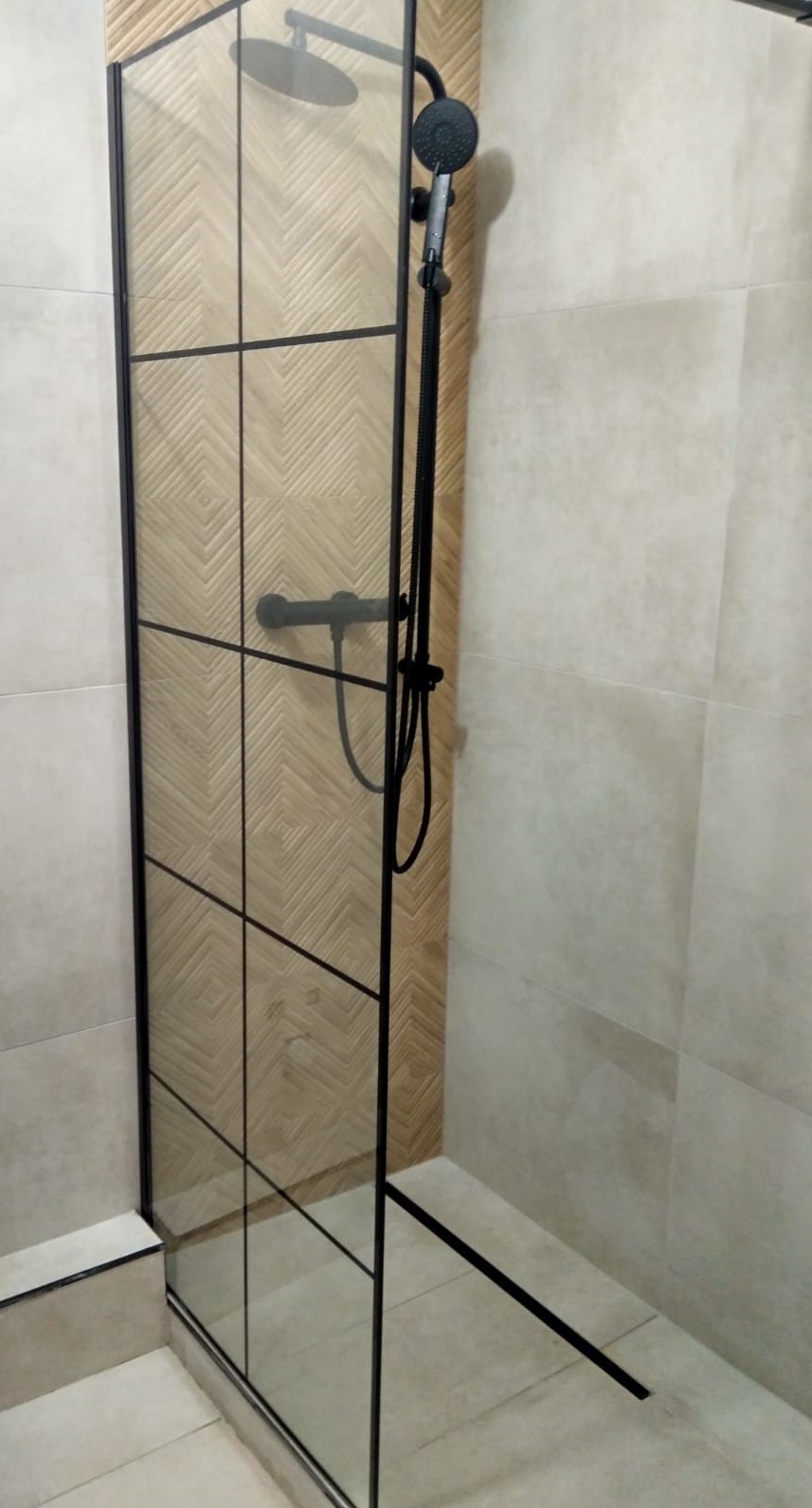 Kabina prysznicowa typu walk-in z czarnymi profilami, deszczownicą i baterią w kolorze czarnym mat, na ścianie płytki imitujące drewno ułożone w jodełkę.