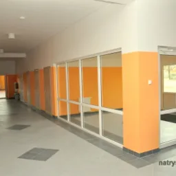 Korytarz biurowy po remoncie: pomarańczowe ściany, szare płytki podłogowe. Widoczne drzwi, okna i znak ewakuacyjny. Jasne, przestronne wnętrze.