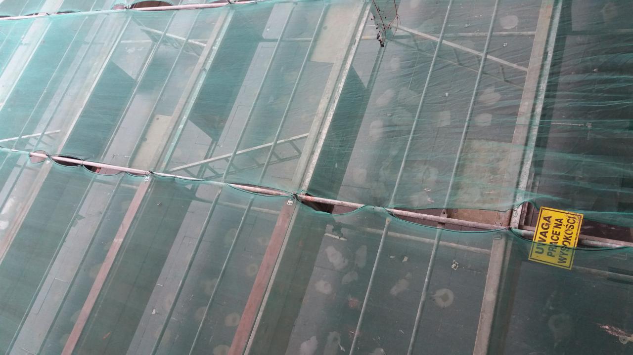 Elewacja budynku w trakcie prac ociepleniowych, zabezpieczona siatką ochronną i rusztowaniami. Żółta tabliczka ostrzegawcza 'Uwaga! Prace na wysokości'.