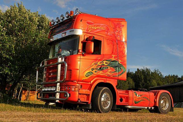 Pomarańczowy ciągnik siodłowy Scania z chromowanym orurowaniem i aerografią przedstawiającą płomienie i postać diabła, zaparkowany na trawiastym terenie.