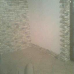 Sak-Bud - Wykończone wnętrze z dekoracyjną ścianą z cegły i fragmentem ściany pomalowanej na biało. Widoczna podłoga z płytek i fragment drzwi.