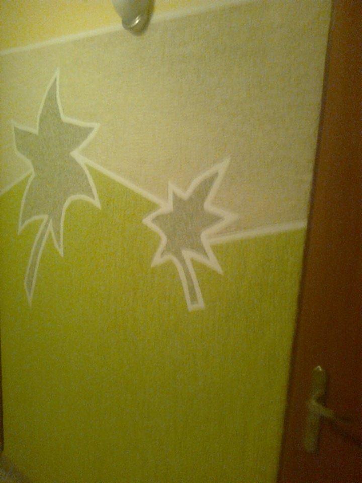 Ściana w pokoju z dwukolorowym malowaniem i motywem liści. Górna część ściany jest jasna, a dolna zielona. Widoczna lampa sufitowa i fragment drzwi.