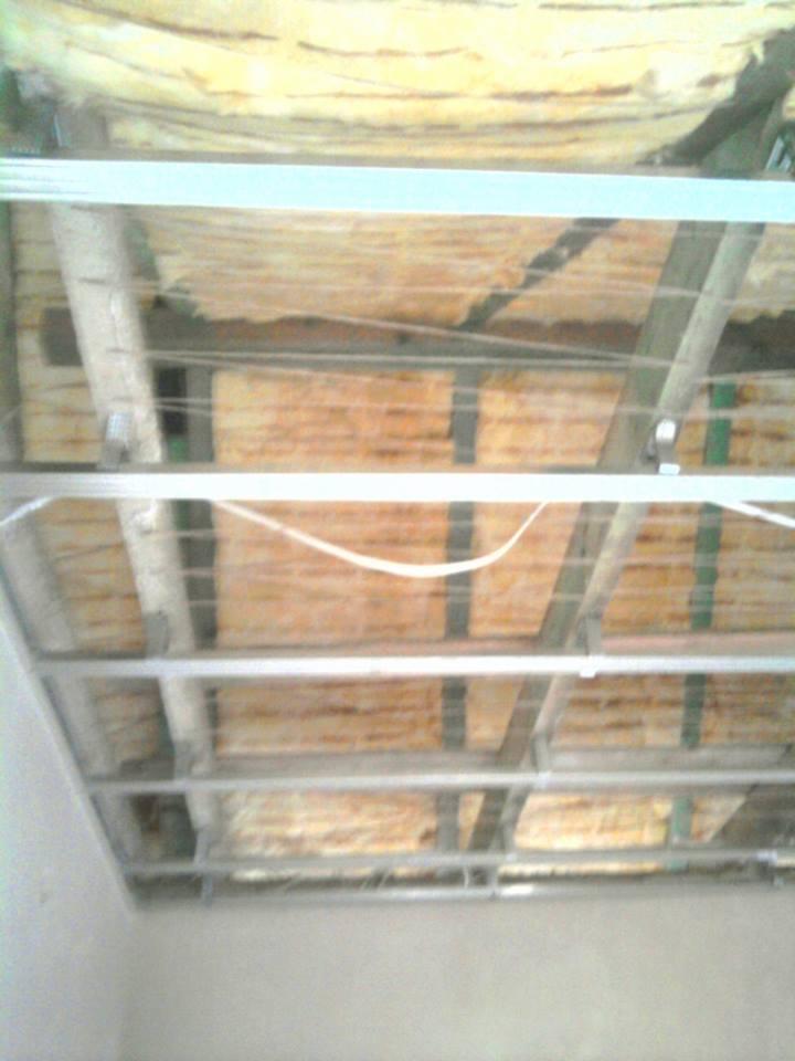 Montaż sufitu podwieszanego: metalowa konstrukcja z izolacją wełną mineralną, widoczne profile i przewody. Prace wykończeniowe w trakcie.