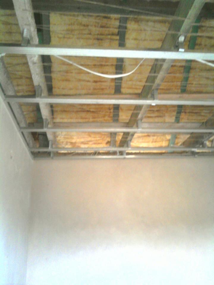 Metalowy stelaż sufitu podwieszanego z izolacją z wełny mineralnej, widok z dołu na przygotowaną konstrukcję przed zakryciem płytami. Ściany przygotowane pod malowanie.