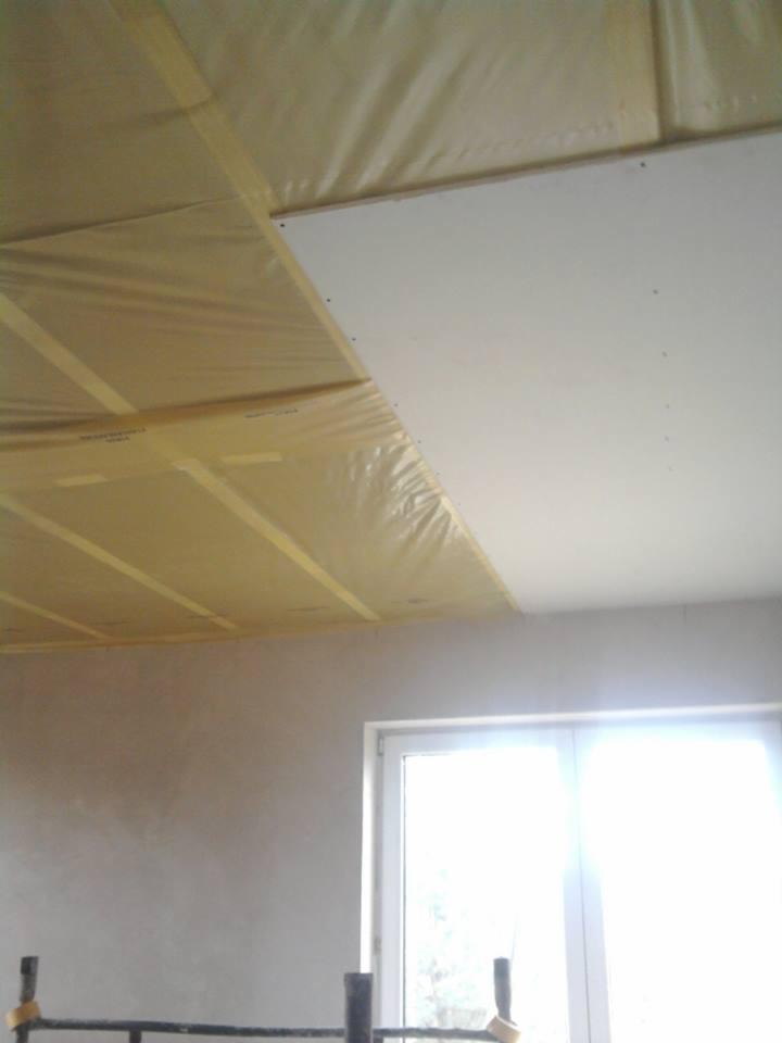 Częściowo zamontowana płyta karton gips na suficie, zabezpieczona folią i taśmą. Widok na okno i fragment ściany w trakcie prac wykończeniowych.