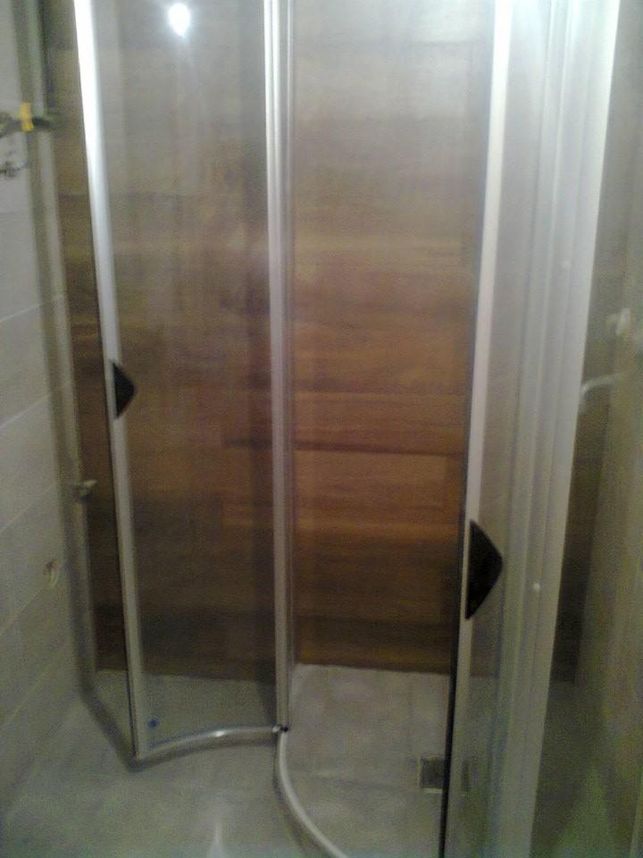 Szklana kabina prysznicowa z drewnianym panelem w tle, narożna, z chromowanymi profilami i czarnymi uchwytami, widok z wnętrza łazienki.