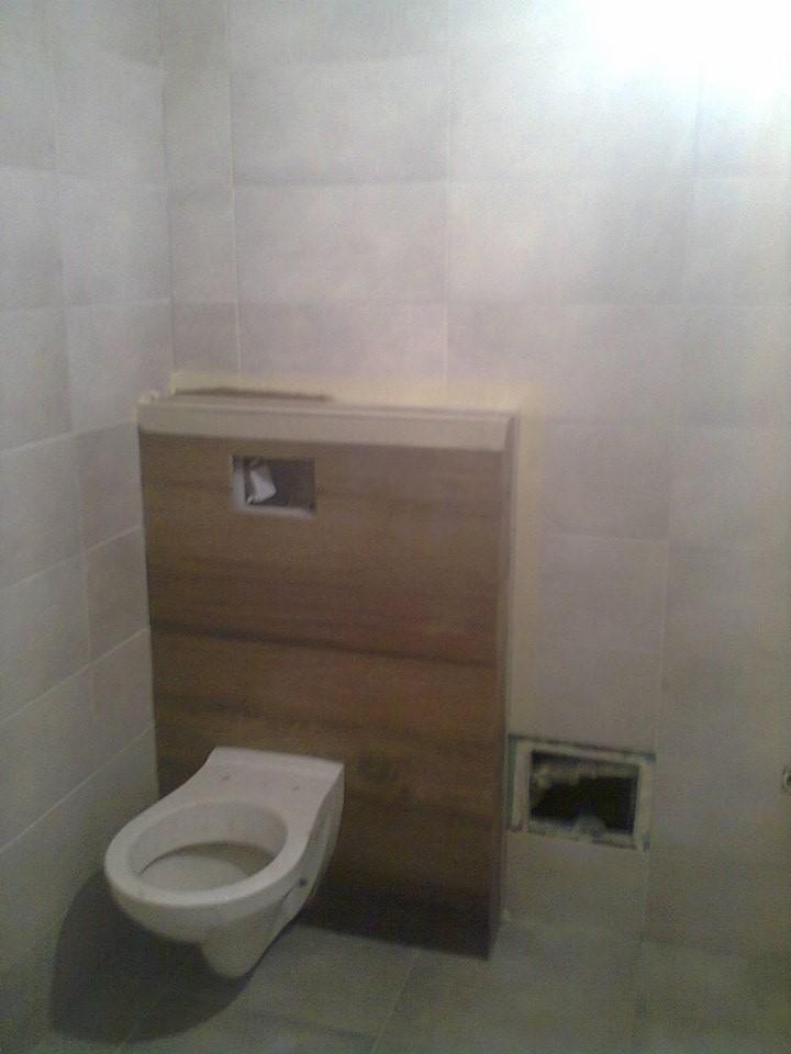 Wisząca toaleta z zabudową w trakcie montażu, widoczne otwory instalacyjne. Szare płytki na ścianach i podłodze. Ujęcie z niskiego kąta, oświetlenie mieszane.