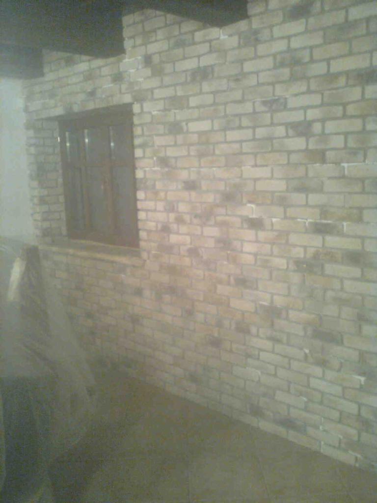 Ściana z cegły dekoracyjnej wewnątrz pomieszczenia z oknem, wykończona w jasnych odcieniach, imitująca stary mur. Widoczny fragment podłogi i przykryte meble.