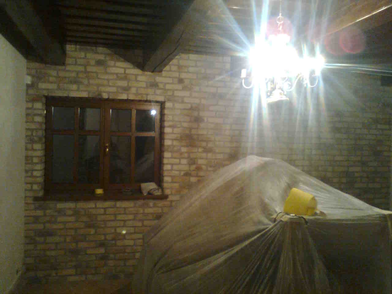 Pokój w trakcie przygotowania do prac remontowych: okno w drewnianej ramie, ściana z cegły, meble zakryte folią ochronną, widoczne drewniane elementy stropu.