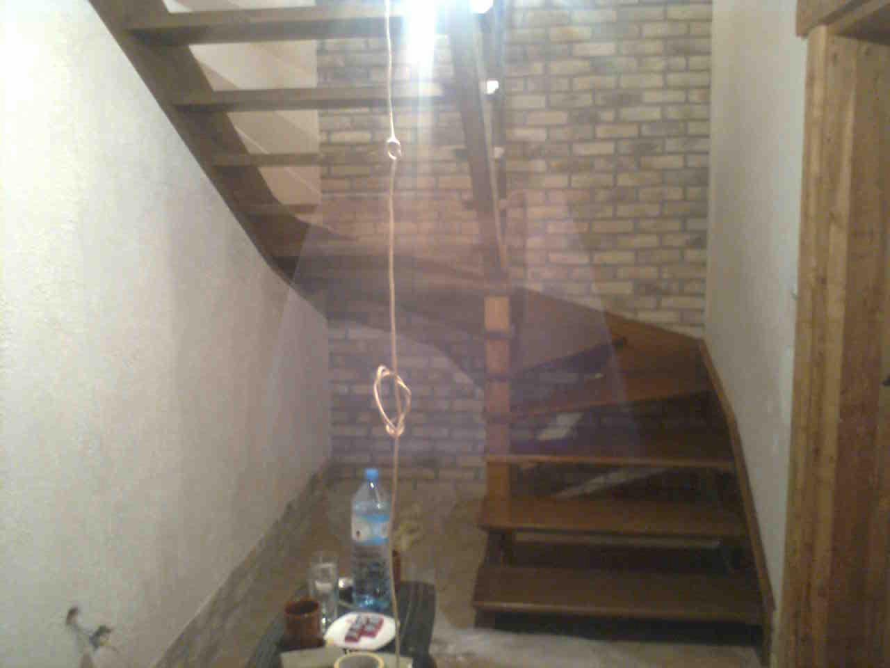 Drewniane schody w jasnym wnętrzu z ceglaną ścianą i gładzią, oświetlone wiszącą żarówką, z detalami jak butelka wody i naczynia na stole. Ujęcie z dołu schodów.