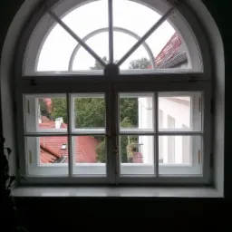 Białe, łukowe okno z podziałami, widok na zewnątrz z zielenią i czerwonym dachem. Oświetlenie dzienne, kontrastowe cienie w pomieszczeniu.