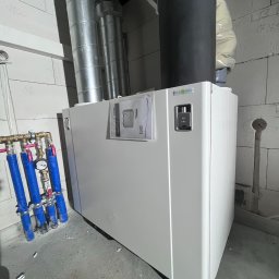 EKO NORD DANIEL ŁAGA - Biała jednostka centralna wentylacyjna Ekonord z podłączonymi rurami wentylacyjnymi i instalacją hydrauliczną, zamontowana przy ścianie z bloczków betonowych.