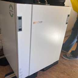 EKO NORD DANIEL ŁAGA - Biała jednostka rekuperacyjna marki Flexit z widocznym logo i naklejką Eko Nord, umieszczona na czarnych podkładkach na podłodze z płyt OSB, z widocznymi elementami instalacji u góry i fragmentem...