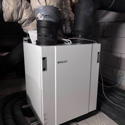 EKO NORD DANIEL ŁAGA - Biała centrala wentylacyjna Flexit z podłączonymi przewodami wentylacyjnymi w surowym pomieszczeniu technicznym.