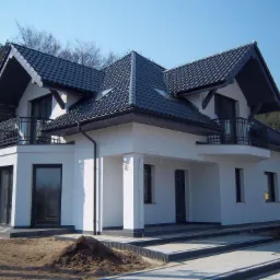 Nowoczesny, biały dom z ciemną dachówką i balkonami, otoczony zielenią i z wykończonym podjazdem.