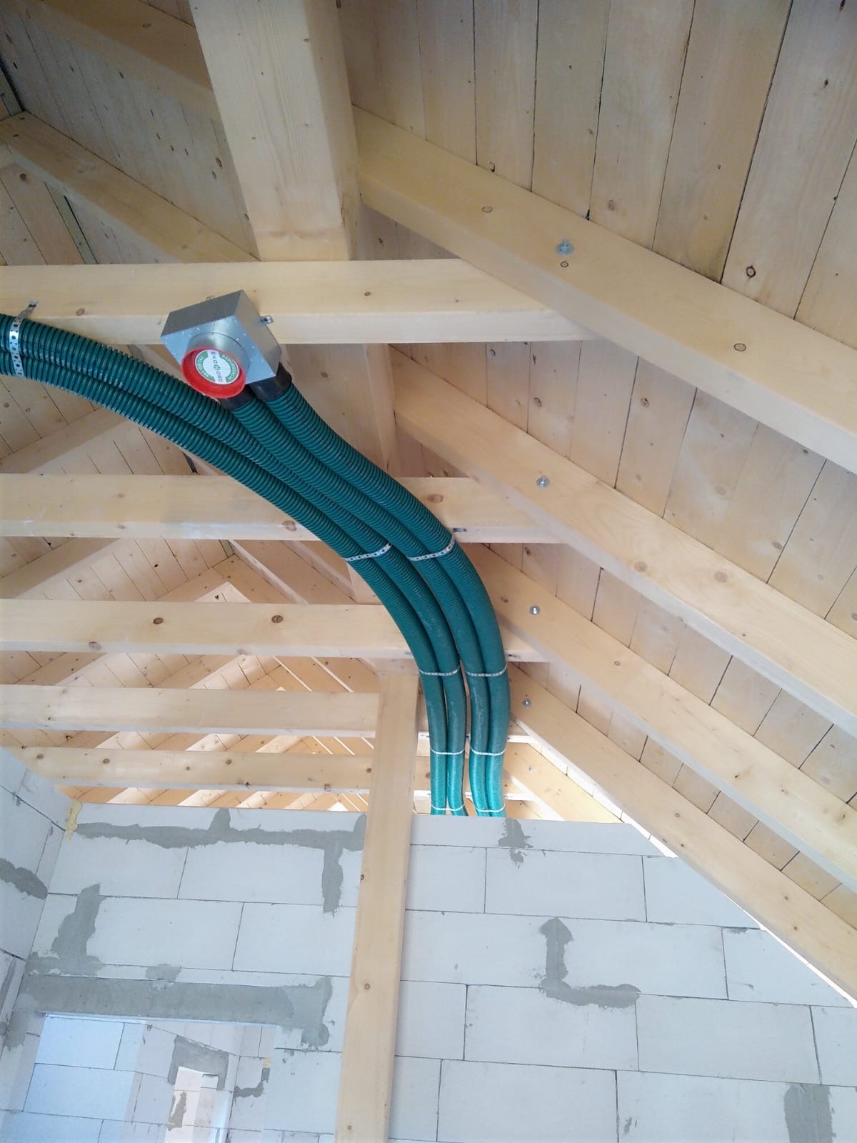 Trzy zielone, karbowane rury wentylacyjne przymocowane do drewnianej konstrukcji dachu w surowym budynku z białych bloczków, widoczny srebrny rozdzielacz z czerwonym elementem.