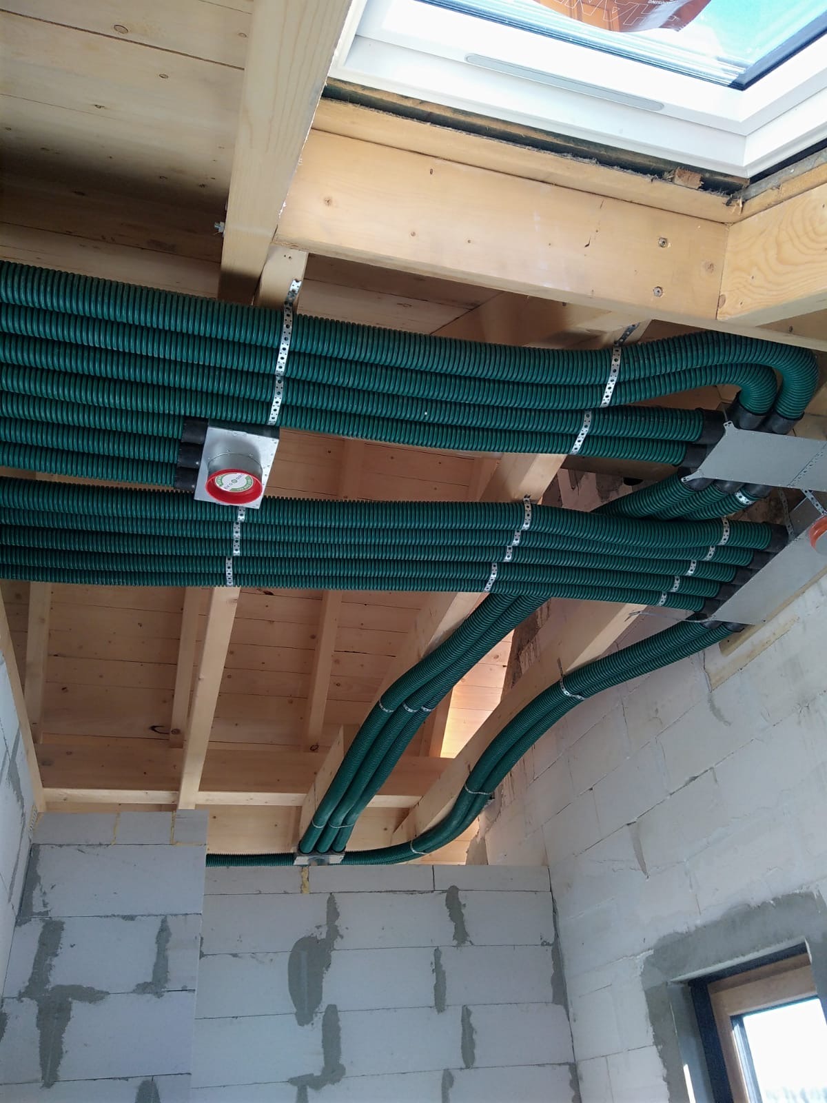 Zielone, karbowane rury wentylacyjne zamontowane pod drewnianym sufitem, przymocowane metalowymi obejmami do belek. Widoczne fragmenty białych ścian z betonu komórkowego i okno dachowe.