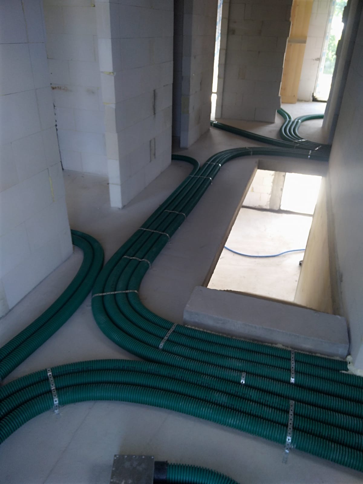 Wnętrze w trakcie budowy z widocznymi, zielonymi przewodami wentylacyjnymi ułożonymi na podłodze i przymocowanymi metalowymi obejmami. Przewody rozchodzą się w różnych kierunkach, prowadząc...