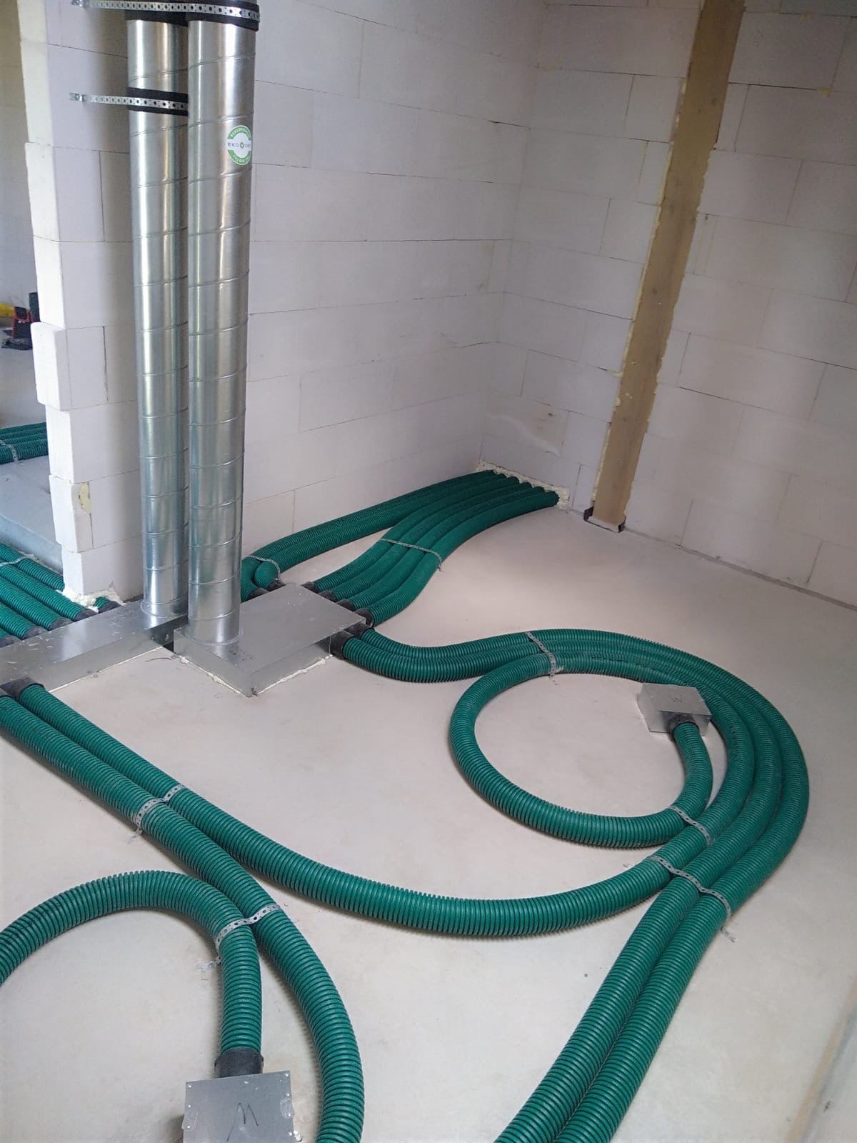 Surowe wnętrze z widocznymi elementami instalacji wentylacyjnej: metalowe rury pionowe i zielone, elastyczne przewody poziome, zamocowane do podłoża w regularnych odstępach.